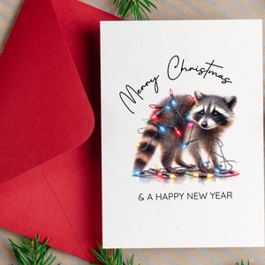 以下が含まれることがあります： カラフルな電飾をまとったアライグマが描かれたクリスマスカード。「Merry Christmas & A Happy New Year」の文字。左側には赤い封筒が見える。