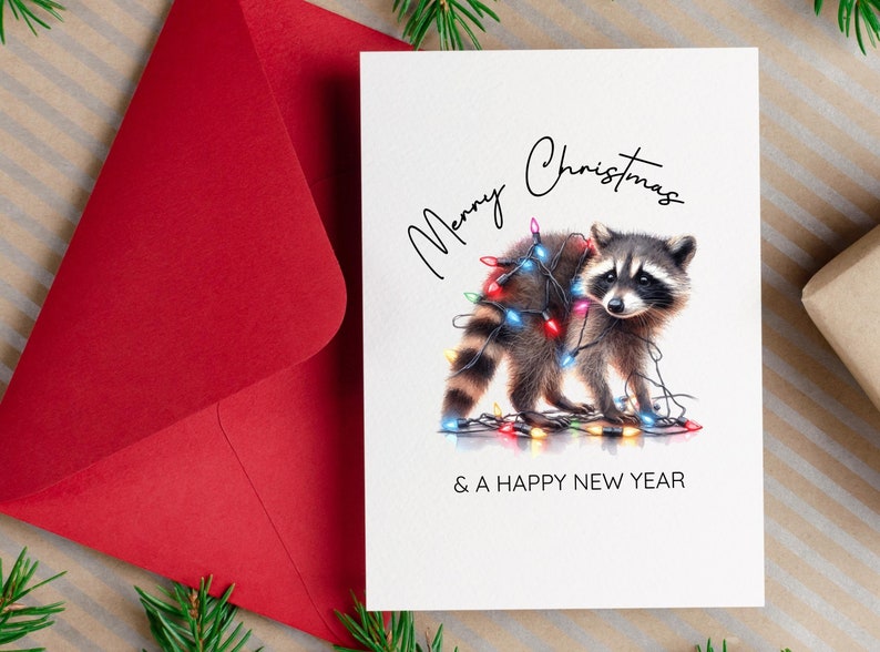 以下が含まれることがあります： カラフルな電飾をまとったアライグマが描かれたクリスマスカード。「Merry Christmas & A Happy New Year」の文字。左側には赤い封筒が見える。