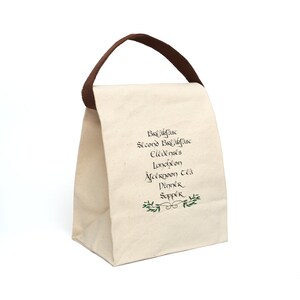 Può includere: Una borsa da pranzo in tela bianca con manici marroni. La borsa ha un design verde e nero con le parole "Colazione, Seconda colazione, Elevenses, Pranzo, Merenda, Cena, Cena".