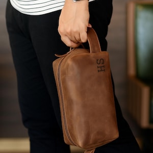 May include: A brown leather wash bag with a handle and zip closure. The initials "HS" are engraved on the front. The person is wearing a watch with a dark face and a black and white striped top.