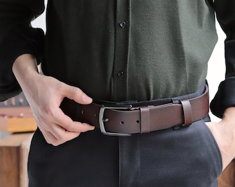May include: A brown leather belt with a silver buckle. The belt is worn over a dark green shirt and dark brown pants.