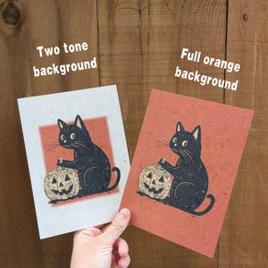 May include: Two Halloween-themed prints featuring a black cat holding a carved pumpkin. One print has a two-tone background, the other a full orange background. The prints are held against a wooden background with a spiderweb.