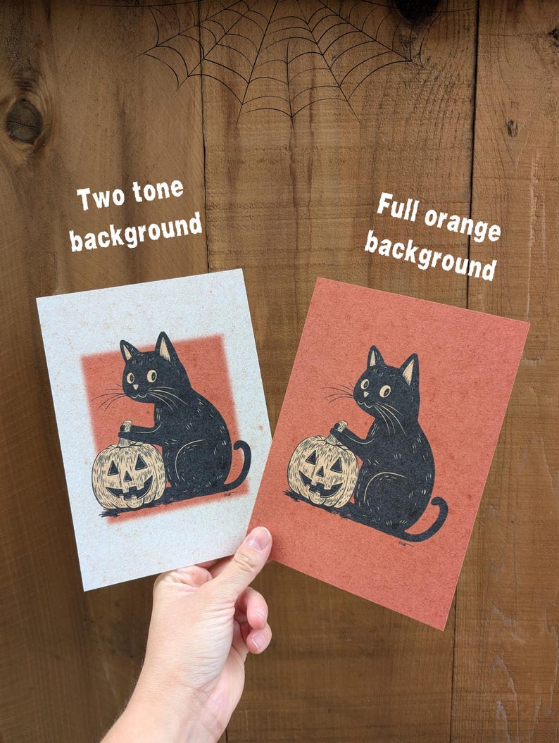 May include: Two Halloween-themed prints featuring a black cat holding a carved pumpkin. One print has a two-tone background, the other a full orange background. The prints are held against a wooden background with a spiderweb.