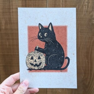 May include: A black cat illustration with a carved pumpkin. The cat is seated, holding a carving tool, with the pumpkin in front. The artwork is on textured paper with a red-orange square background, perfect for Halloween.