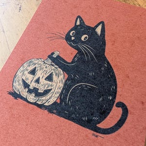 May include: A black cat illustration with a carved pumpkin on a reddish-orange background. The cat is sitting and appears to be holding the pumpkin. The pumpkin has a carved face. The artwork has a vintage, hand-drawn style.