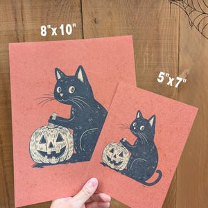 May include: Two prints featuring a black cat holding a carved pumpkin. The prints are on a reddish-orange background. The larger print measures 8" x 10", and the smaller one is 5" x 7". The cat and pumpkin are in a vintage style.