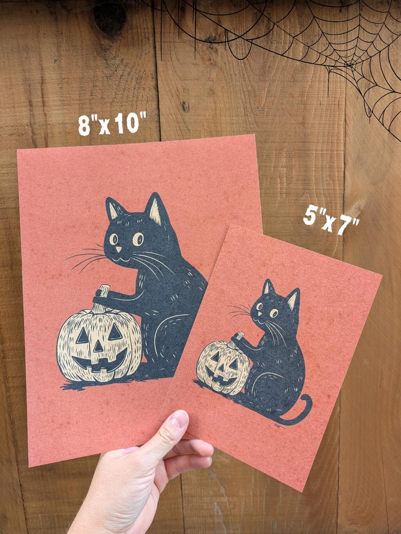 May include: Two prints featuring a black cat holding a carved pumpkin. The prints are on a reddish-orange background. The larger print measures 8" x 10", and the smaller one is 5" x 7". The cat and pumpkin are in a vintage style.