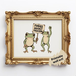 May include: Printable wall art featuring two green frogs. One frog holds a sign that reads "Frogs Against Fascism," while the other raises a fist. The artwork is framed in a gold ornate frame.