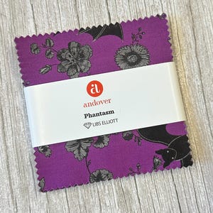 May include: A stack of purple fabric squares with a black floral and bat pattern. The fabric is wrapped with a white band that reads "andover Phantasm LIBS ELLIOTT". The fabric has a scalloped edge.