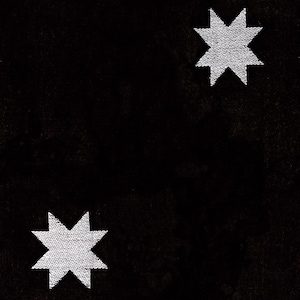 May include: A solid black fabric background featuring two white embroidered eight-pointed stars. The stars are positioned diagonally across the frame, with one located in the upper right corner and the other in the lower left corner of the textile.