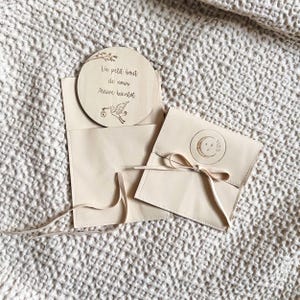 May include: A light beige baby announcement set. The set includes a round wooden announcement with text and a stork illustration, and two matching beige envelopes. One envelope is tied with a ribbon.