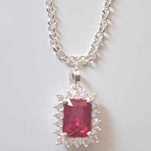 May include: A silver necklace with a rectangular, ruby-colored pendant. The pendant is surrounded by a halo of smaller, clear stones. The chain is a linked design, and the necklace is displayed against a white background.