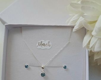 May include: A white gift box with a silver necklace and earrings. The necklace has a small blue gemstone pendant. The box has the word "March" written on it.