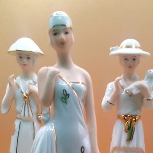 May include: Three porcelain figurines of women in elegant gowns and hats. The figures are primarily white with gold accents and delicate floral details. One figure holds a large leaf-shaped object.