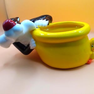 May include: A ceramic planter featuring Sylvester the Cat and Tweety Bird. Sylvester is perched on the side, with a red nose and a black and white body. Tweety Bird is on the other side. The planter is yellow.