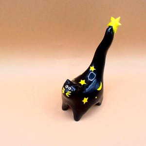 May include: A black ceramic cat figurine with yellow star and moon accents. The cat is reaching up to a yellow star. The figurine is on a light brown surface.