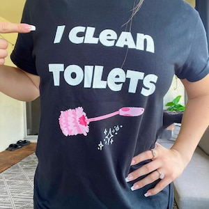 May include: A black short-sleeved t-shirt featuring the white text "I CLEAN TOILETS" printed on the chest. Below the text is a graphic of a pink toilet brush with a long handle, accompanied by small white star-shaped sparkle icons.