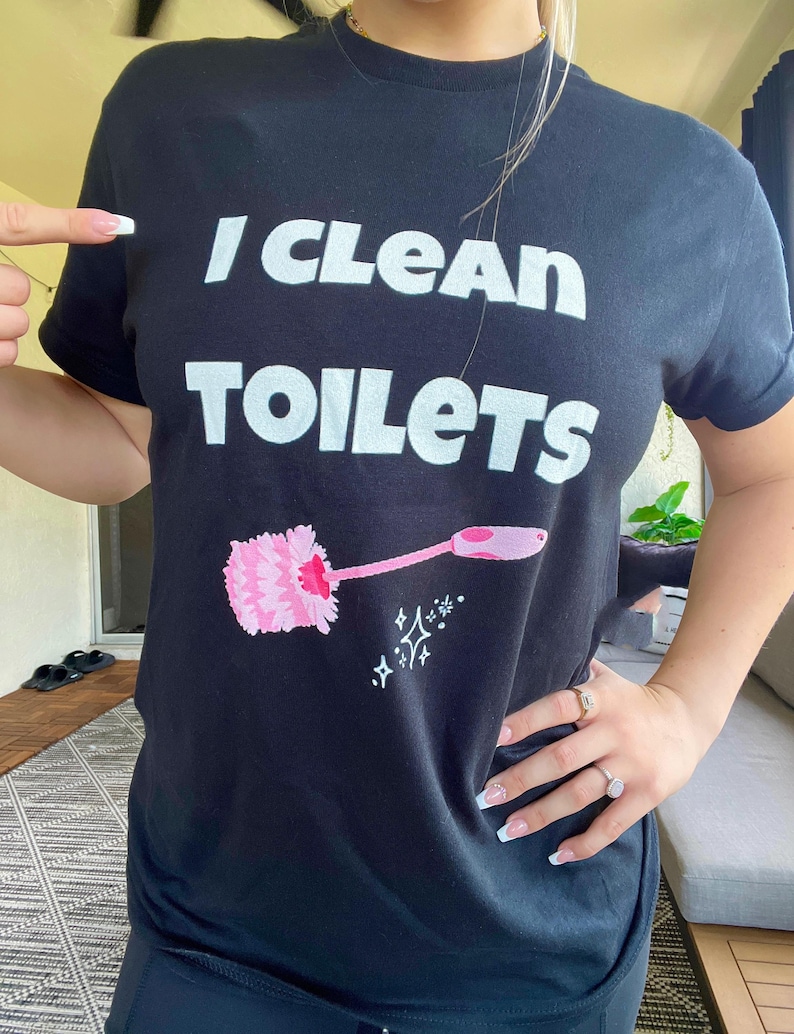 May include: A black short-sleeved t-shirt featuring the white text "I CLEAN TOILETS" printed on the chest. Below the text is a graphic of a pink toilet brush with a long handle, accompanied by small white star-shaped sparkle icons.