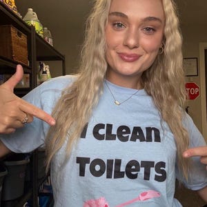 May include: A light blue short-sleeved t-shirt featuring the black text "I CLEAN TOILETS" printed on the front. Below the text is a graphic illustration of a pink toilet brush with a pink handle and small black star accents.