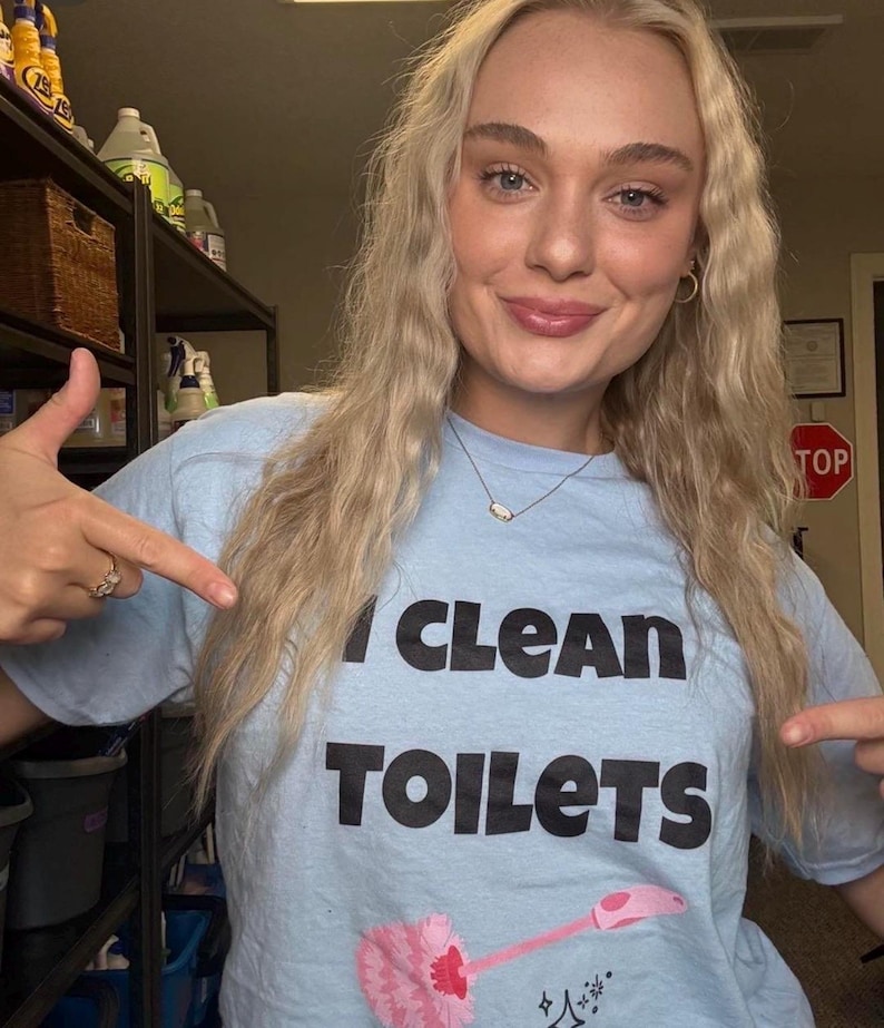 May include: A light blue short-sleeved t-shirt featuring the black text "I CLEAN TOILETS" printed on the front. Below the text is a graphic illustration of a pink toilet brush with a pink handle and small black star accents.