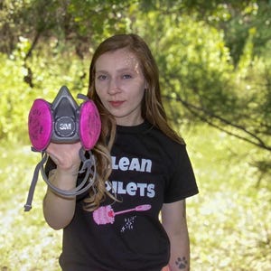 May include: A gray 3M respirator mask with two circular pink filters attached to the sides. The mask features a central circular valve and adjustable gray straps. The filters are clearly labeled with the text 2097 and the 3M brand logo.