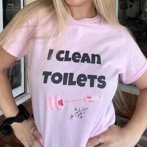 May include: A light pink short-sleeved t-shirt featuring the black text I CLEAN TOILETS printed on the chest. Below the text is a graphic illustration of a pink toilet brush and a small black sparkle icon centered on the front of the garment.