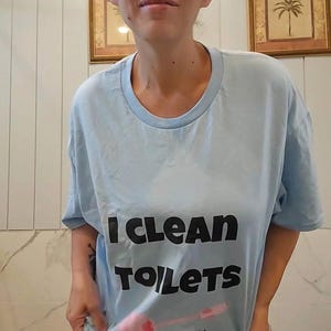 May include: A light blue short-sleeved t-shirt featuring the black text I CLEAN TOILETS and a graphic of a pink toilet brush. The shirt is paired with multicolored patterned leggings featuring shades of blue, pink, and white in a floral design.