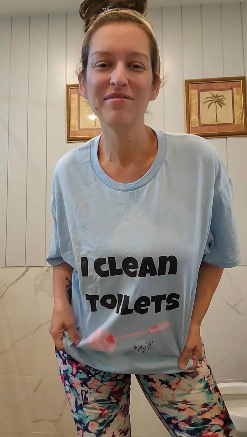 May include: A light blue short-sleeved t-shirt featuring the black text I CLEAN TOILETS and a graphic of a pink toilet brush. The shirt is paired with multicolored patterned leggings featuring shades of blue, pink, and white in a floral design.