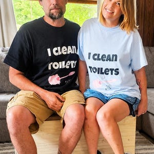 May include: A black t-shirt and a light blue t-shirt, both featuring the text "I CLEAN TOILETS" in bold black lettering above a graphic of a pink toilet brush. The shirts are short-sleeved with a crew neckline, made of a standard cotton fabric.