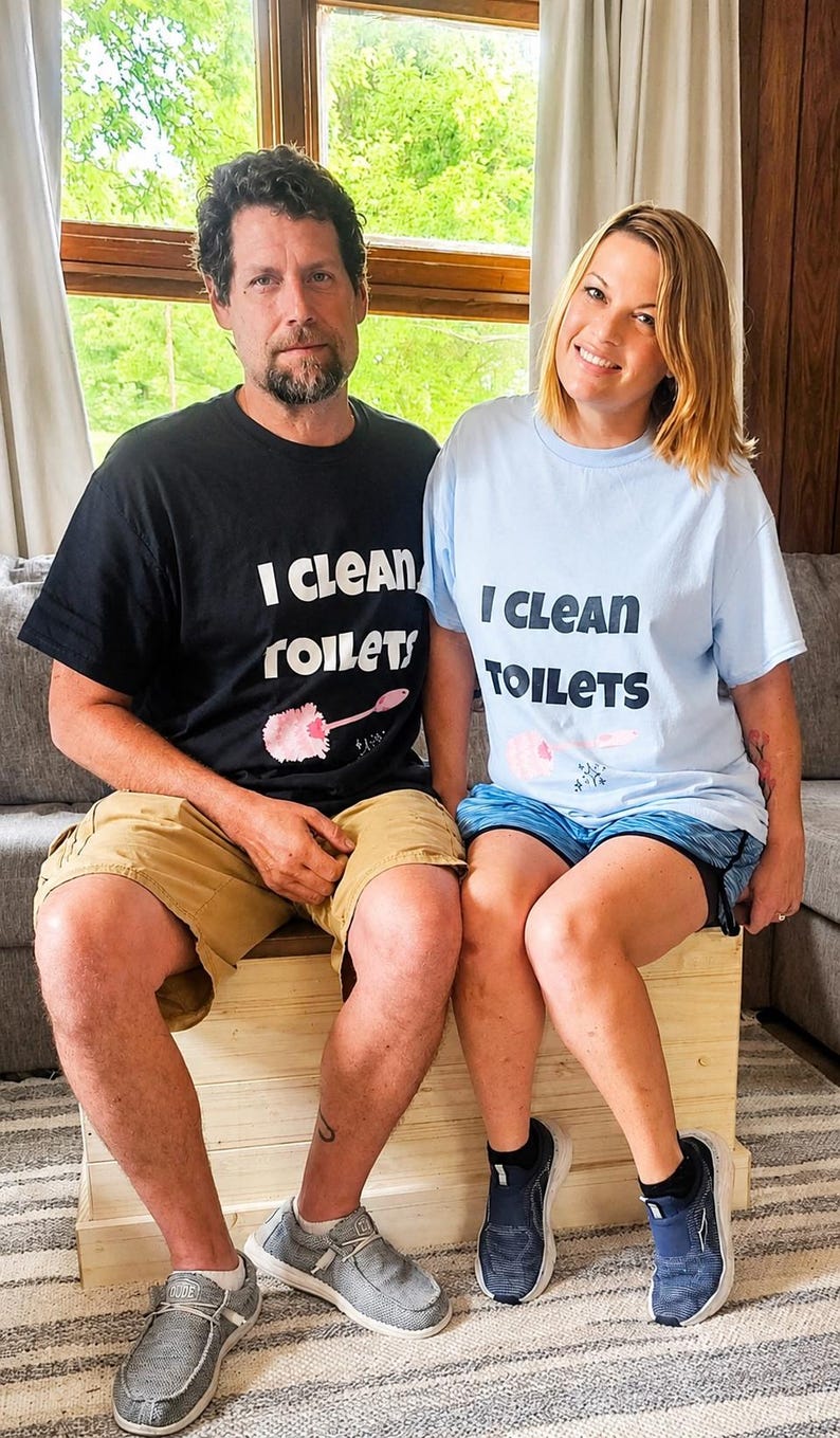 May include: A black t-shirt and a light blue t-shirt, both featuring the text "I CLEAN TOILETS" in bold black lettering above a graphic of a pink toilet brush. The shirts are short-sleeved with a crew neckline, made of a standard cotton fabric.