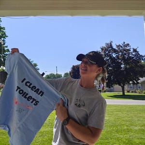 May include: A light blue t-shirt with the text I CLEAN TOILETS printed in black, featuring a graphic of a toilet brush. The shirt is held up alongside a grey t-shirt with a small circular logo on the chest and a black baseball cap with gold embroidery.