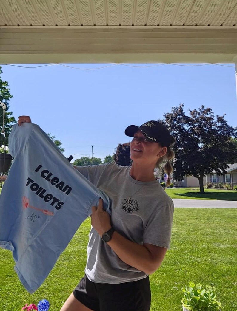 May include: A light blue t-shirt with the text I CLEAN TOILETS printed in black, featuring a graphic of a toilet brush. The shirt is held up alongside a grey t-shirt with a small circular logo on the chest and a black baseball cap with gold embroidery.