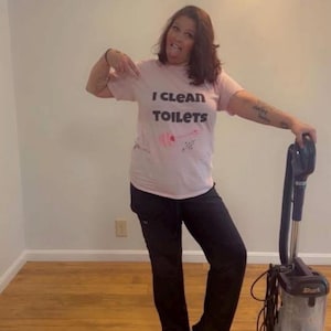 May include: A light pink short-sleeved t-shirt with the black text I CLEAN TOILETS and a small pink graphic of a toilet brush. The shirt is paired with black trousers and is positioned next to a dark gray and black upright Shark vacuum cleaner.