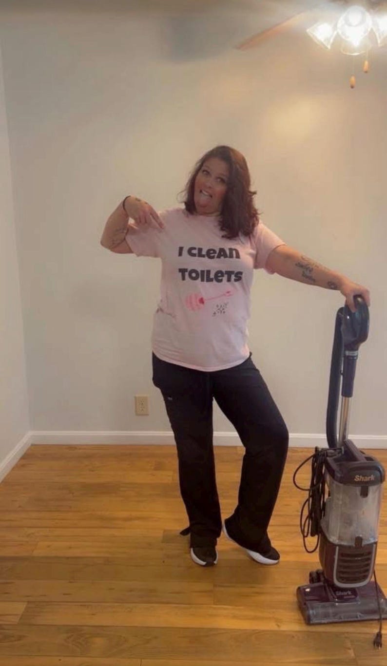 May include: A light pink short-sleeved t-shirt with the black text I CLEAN TOILETS and a small pink graphic of a toilet brush. The shirt is paired with black trousers and is positioned next to a dark gray and black upright Shark vacuum cleaner.