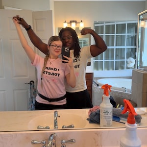 May include: A light pink short-sleeved t-shirt featuring the black text I CLEAN TOILETS and a small pink graphic. The shirt has a crew neckline and is worn with a black belt. The text is mirrored in the reflection of the bathroom mirror.