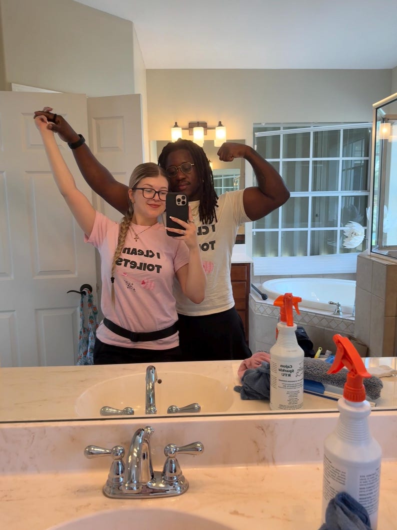 May include: A light pink short-sleeved t-shirt featuring the black text I CLEAN TOILETS and a small pink graphic. The shirt has a crew neckline and is worn with a black belt. The text is mirrored in the reflection of the bathroom mirror.