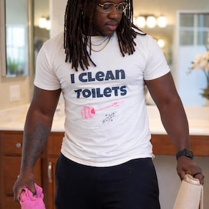 May include: A white short-sleeved t-shirt featuring the text "I CLEAN TOILETS" in dark blue letters, accompanied by a graphic of a pink toilet brush and small sparkle icons. The shirt is paired with black pants, a pink cloth, and a beige mat.