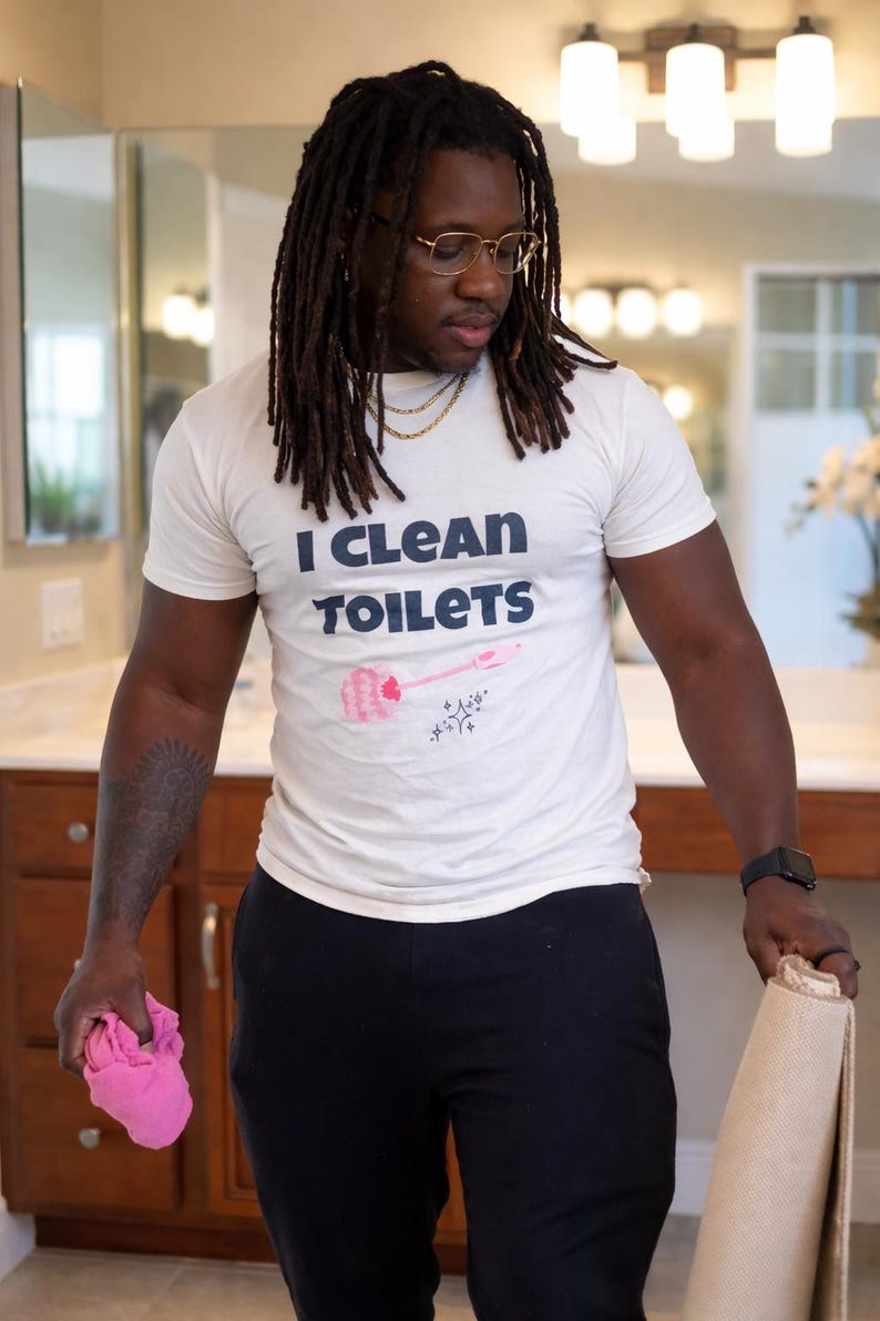 May include: A white short-sleeved t-shirt featuring the text "I CLEAN TOILETS" in dark blue letters, accompanied by a graphic of a pink toilet brush and small sparkle icons. The shirt is paired with black pants, a pink cloth, and a beige mat.