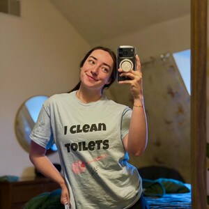 May include: A light blue short-sleeved t-shirt featuring the black text "I CLEAN TOILETS" printed on the front. Below the text is a graphic illustration of a pink toilet brush with a small black star accent.