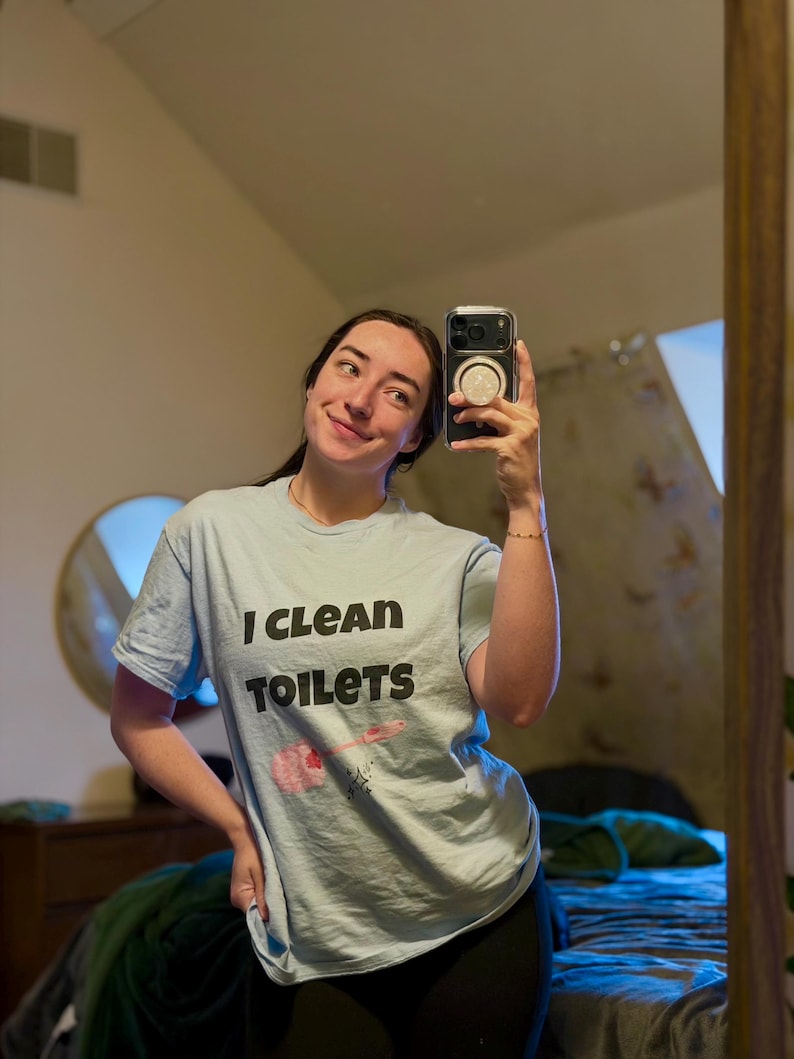 May include: A light blue short-sleeved t-shirt featuring the black text "I CLEAN TOILETS" printed on the front. Below the text is a graphic illustration of a pink toilet brush with a small black star accent.
