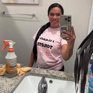 May include: A light pink short-sleeved t-shirt featuring the black text I CLEAN TOILETS and a graphic of a pink toilet brush with small star accents. The shirt is made of a cotton-like fabric and has a crew neckline and a standard fit.