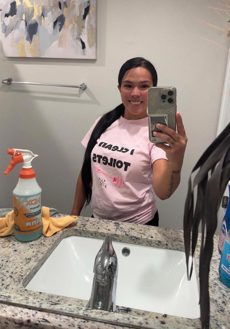 May include: A light pink short-sleeved t-shirt featuring the black text I CLEAN TOILETS and a graphic of a pink toilet brush with small star accents. The shirt is made of a cotton-like fabric and has a crew neckline and a standard fit.