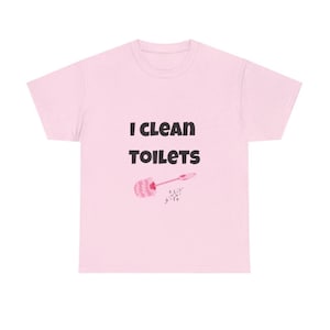 May include: A light pink short-sleeved t-shirt featuring the black text "I CLEAN TOILETS" printed on the front. Below the text is a small graphic of a pink toilet brush and a tiny sparkling star icon, centered on the chest of the garment.