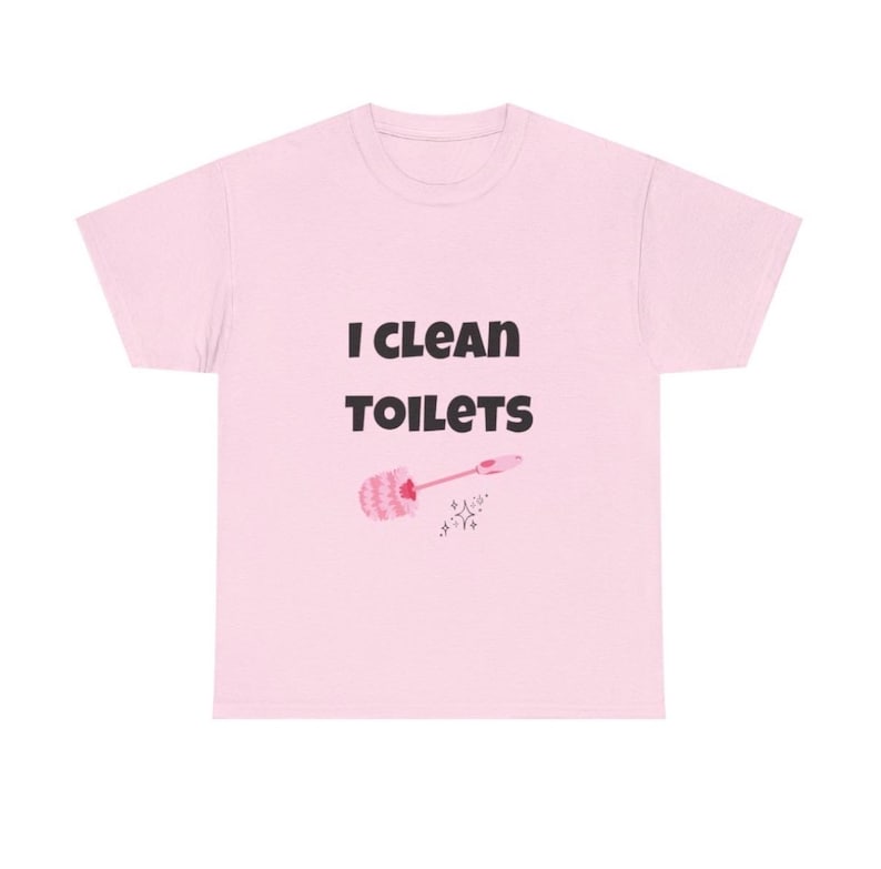 May include: A light pink short-sleeved t-shirt featuring the black text "I CLEAN TOILETS" printed on the front. Below the text is a small graphic of a pink toilet brush and a tiny sparkling star icon, centered on the chest of the garment.