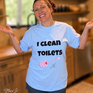 May include: A light blue short-sleeve t-shirt featuring the black text I CLEAN TOILETS printed on the front. Below the text is a graphic of a pink toilet brush with a white brush head and small black sparkle icons surrounding the handle.