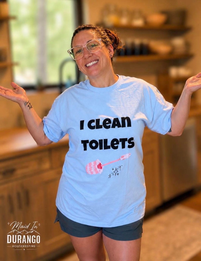 May include: A light blue short-sleeve t-shirt featuring the black text I CLEAN TOILETS printed on the front. Below the text is a graphic of a pink toilet brush with a white brush head and small black sparkle icons surrounding the handle.