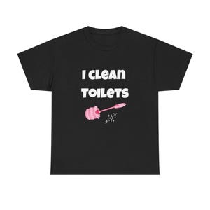 May include: A black short-sleeve t-shirt featuring the white text "I CLEAN TOILETS" printed on the chest. Below the text is a graphic of a pink toilet brush with a fluffy head and a small white sparkle icon to the right of the brush handle.