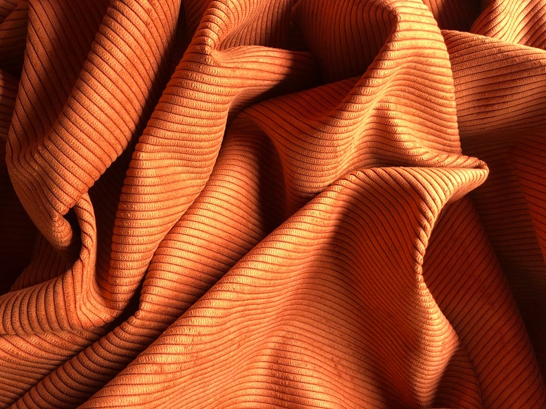 May include: Close-up of a rust-colored corduroy fabric with a ribbed texture. The fabric is folded and draped, creating a soft, flowing look.