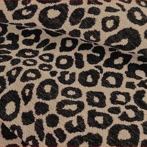 Pode incluir: Um tecido têxtil com um padrão de leopardo repetitivo com manchas pretas sobre um fundo bege claro. O material tem uma superfície texturizada com fibras tecidas visíveis e dobras que criam sombras subtis em todo o design.