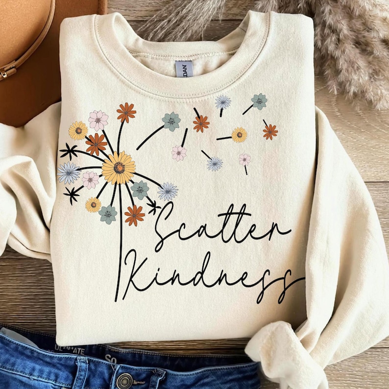 May include: Cream-colored sweatshirt featuring a dandelion design with various colored flowers and the words "Scatter Kindness" in a handwritten font. The sweatshirt is folded, with a portion of blue jeans visible.